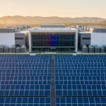 Expansive rows of blue solar panels leading to a sleek data center with cooling units and a glass-walled server hall faintly glowing with RGB light, lit by warm golden-hour sun, with distant power lines and low hills softly blurred in the background.
