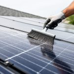 Close-up of dusty solar panels on a residential rooftop while a soft-bristle brush and squeegee clean the panel surface.